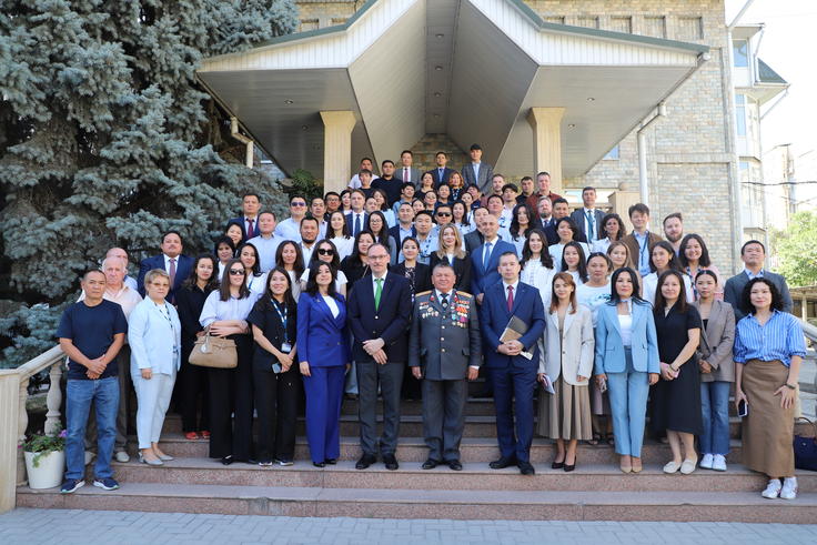 Large group of people posing on steps outside a building, some in suits and uniforms, with greenery in the background.